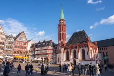 Frankfurt, Almanya 'daki Romerberg Meydanı' nda Eski Aziz Niklas Kilisesi, yarı ahşap binalar ve mavi gökyüzünün altındaki canlı atmosferin tadını çıkaran insanlar yer alıyor..