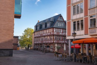 Frankfurt, Almanya 'da bir kaldırım taşı caddesi, kırmızı ve beyaz yarı ahşap bir bina, turuncu şemsiyelerin altında açık hava koltukları ve yakınlardaki modern binalar..