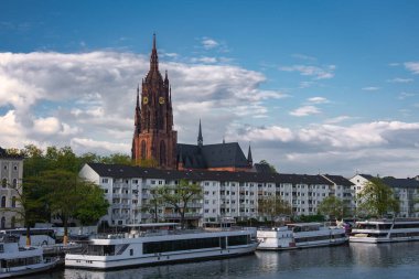 Frankfurt Katedrali 'nin Gotik kulesi parçalı bulutlu bir gökyüzüne karşı yükseliyor, Ana Nehir, nehir botları, konut binaları ve yeşillik manzaralı.