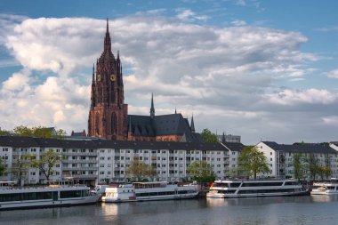 Frankfurt Katedrali 'nin Gotik kulesi bulutlu bir gökyüzüne karşı yükselir, Ana Nehir, rıhtım tekneleri ve ön plandaki modern yerleşim binaları ile.