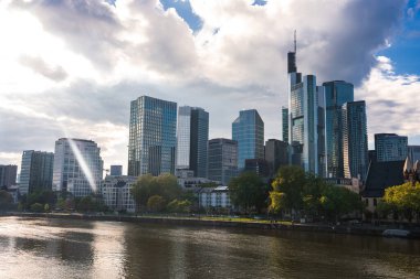 Frankfurt 'un siluetinde Commerzbank Kulesi ve Ana Nehir boyunca cam gökdelenler, yansımalar, yeşillik ve parçalı bulutlu gökyüzü yer almaktadır..