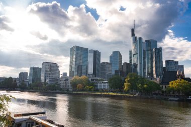 Frankfurt 'un siluetinde Commerzbank Kulesi, cam gökdelenler ve bir kilise yer alır. Ana nehir binaları, ağaçları ve parçalı bulutlu gökyüzünü yansıtır..