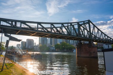 Eiserner Steg köprüsü, Frankfurt, Almanya 'daki Main River' ı demir iskelesi, modern silueti ve sıcak güneş ışığıyla kenetlenmiş bir teknesi ile kaplar..