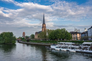 Dreikonigskirche 'nin yüksek kulesi Frankfurt' taki ana nehrin kıyısından yükseliyor, ağaçlarla çevrili, bir gezinti güvertesi, kenetlenmiş tekneler ve karışık mimariyle..