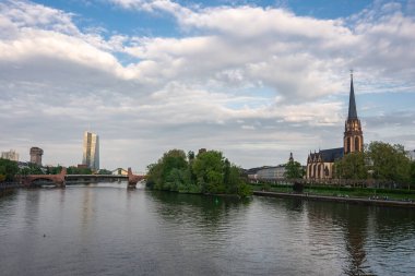 Frankfurt 'un ana nehri Dreikonigskirche kulesini, tarihi bir taş köprüyü ve parçalı bulutlu gökyüzünün altındaki modern ECB kulesini geçer..