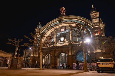 Almanya 'daki Frankfurt Hauptbahnhof' un gece görüşü, aydınlık cephesi, süslü heykelleri, kubbeli kuleleri, çıplak ağaçları, bir taksiyi ve girişe yakın insanları içeriyor..