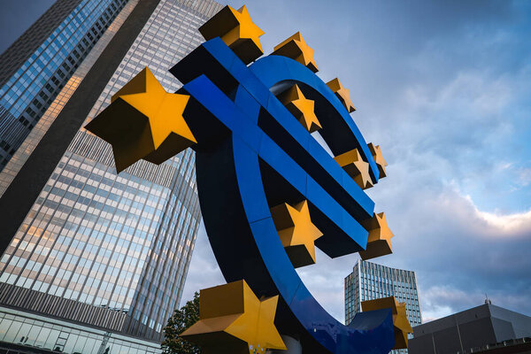 Знаменитая скульптура Евро с желтыми звездами стоит перед современными высотными зданиями, включая бывшую штаб-квартиру Европейского центрального банка, под облачным небом..