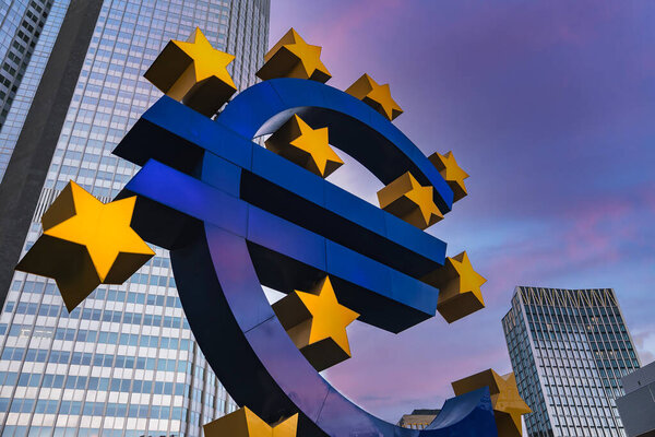 Скульптура евро стоит во Франкфурте, Германия, в окружении небоскребов, включая бывшую штаб-квартиру Европейского центрального банка, под облачным небом.