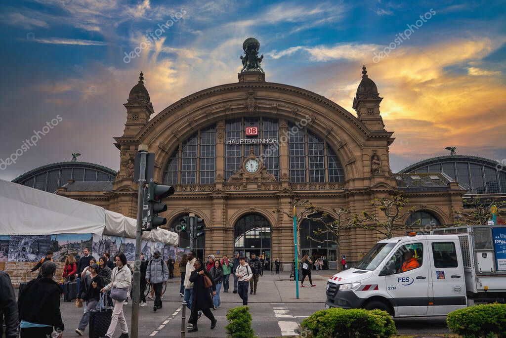 The grand facade of Frankfurt Hauptbahnhof in Germany features a large arched entrance, a clock, and the DB logo, with pedestrians and a white van nearby.