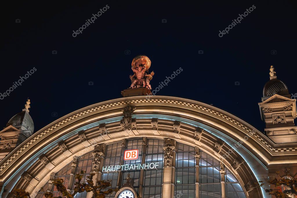 Frankfurt Hauptbahnhof's arched facade glows at night, featuring intricate details, a central statue, and prominent signage under a dark sky.