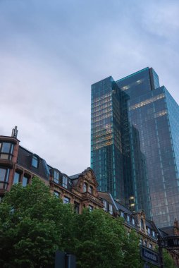Frankfurt 'un alacakaranlıktaki manzarası. Süslü tarihi binaların yanında cam bir gökdelen var. Sokakta ağaçlar ve bulutlu bir akşam gökyüzü var..