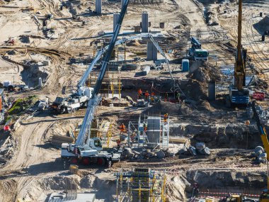 İşçiler, vinçler, beton pompa kamyonu ve iskelesi olan aktif bir inşaat alanı. Özellikler beton sütunları ve düzenli kumlu araziyi güçlendirdi.