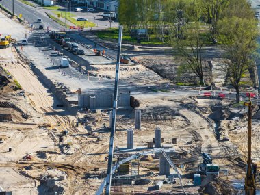 Ağır makineler, işçiler, beton sütunlar ve ağaçlar ve kentsel yapılarla çevrili kısmen inşa edilmiş bir yol ile aktif bir inşaat sahası..