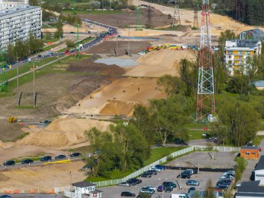 Kum tepecikleri, araçlar, işçiler, kırmızı ve beyaz kule, konut binaları, otopark, yol trafiği ve uzak orman trafiği olan aktif inşaat alanı.