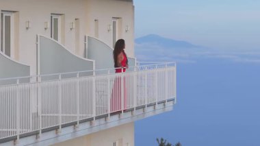 Kırmızı elbiseli bir kadın Amalfi sahil şeridine bakan beyaz korkuluklu bir otel balkonunda duruyor. İnce hareketler onun değişken duruşunu gösteriyor..