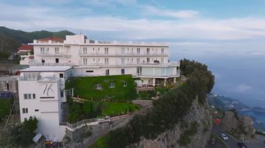 Amalfi kıyı şeridi yakınlarındaki yeşil, engebeli uçurumlar, yollar ve kırmızı çatılı evlerle çevrili bir uçurum üzerindeki beyaz çok katlı otelin havadan görüntüsü..