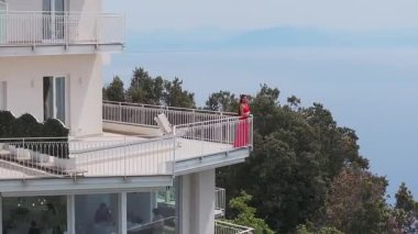 Amalfi kıyısındaki modern beyaz bir otelde teraslar, balkonda kırmızılı bir kadın, cam duvarlı bir yemek alanı ve yemyeşil bir çevre bulunmaktadır..