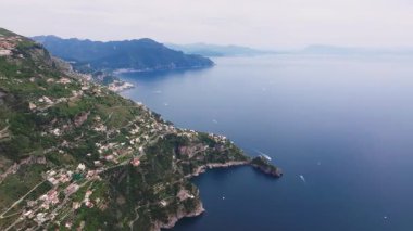 İtalya 'daki Amalfi kıyı şeridinin sakin hava manzarası, kayalık bir yarımadada beyaz binalar, hareket halindeki tekneler ve hafif bulutlu bir gökyüzü..