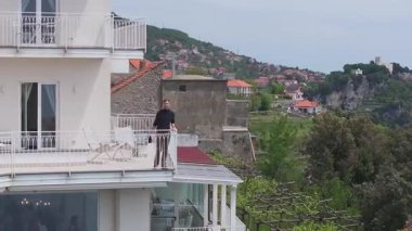 Lüks bir otelin balkonunda beyaz bir cephe ve dekoratif panellerle yürüyen bir adam. Yeşillik ve kıyı manzaraları arka planda görülebilir.