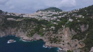 İtalya, Capri 'nin havadan görünüşü, kayalıkları, beyaz binaları, yemyeşil alanları ve kayalık çıkıntıları gösteriyor. Kıyı şeridi boyunca bulutlu bir gökyüzünün altında yumuşak bir hareket.
