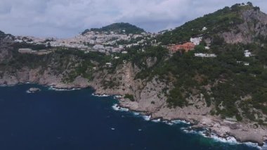 İtalya, Capri 'nin havadan görünüşü, uçurumları, mavi denizi ve yeşil tepelerdeki beyaz binaları gösteriyor. Kamera sahil şeridinde hafif bulutlarla sorunsuzca ilerliyor..