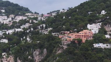 İtalya 'nın Capri kentinin havadan görünüşü yamaçtaki beyaz ve pastel binaları gösteriyor. Kamera kayalıklardan, yeşillikten ve Akdeniz mimarisinden geçiyor..