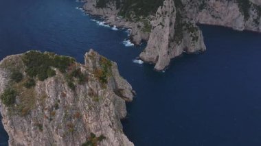 İtalya 'nın Capri kentinin havadan görünüşü, kayalıkları, engebeli kıyı şeritlerini, yamaçlardaki binaları, yemyeşil alanları ve bulutlu gökyüzü altında eşsiz kaya oluşumlarını gösteriyor..