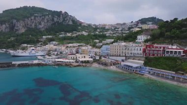 Feribotları, tekneleri, renkli binaları ve yemyeşil tepeleri olan Capri 'nin marinasının havadan görüntüsü. Kamera sahil şeridinde ve yamaçta sorunsuzca hareket ediyor..