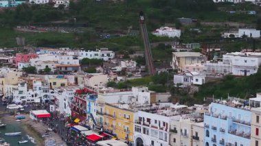 Capri, İtalya 'nın havadan görünüşü, renkli rıhtım binaları, tekneli kalabalık bir marina, füniküler demiryolu ve gezinti güvertesi boyunca insanlar..