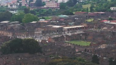 Pompei 'nin İtalya' daki harabelerinin hava manzarasında taş duvarlar, sütunlar, kırmızı kiremitli çatılar, avlular, sokaklar ve çevreleyen yeşillikler yavaş çekimde yer alıyor..