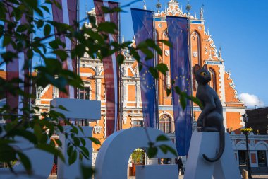 Riga, Letonya 'daki Siyahların Evi' nde kırmızı tuğla cephe, AB ve Letonya bayrakları ve RIGA harflerinin üzerindeki Kedi Heykeli Akımı yer alıyor..
