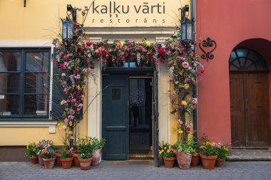 Riga 'nın eski kasabasındaki Kalku Varti restoranının girişinde canlı bir çiçek kemeri, saksı bitkileri, ılık tonlu dış görünüş ve kaldırım taşı caddesi yer alıyor..