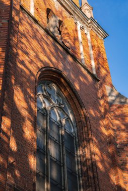 Riga 'nın eski kasabasında kırmızı tuğlalı bir cephede karmaşık Gotik bir iz sürücü ile uzun kemerli bir pencere. Güneş ışığı ve ağaç gölgeleri dinamik desenler oluşturur.