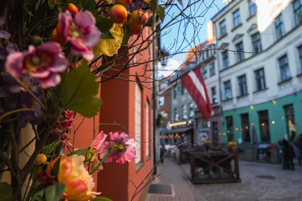 Riga 'nın eski kasabasındaki bir kaldırım taşı caddesinde pastel binalar, bir Letonya bayrağı, renkli çiçekler, küçük kafeler ve rahat bir ortamda sicim ışıkları yer alıyor..