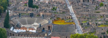 İtalya 'daki Pompei harabelerinin havadan görünüşü, yarı daire şeklinde oturma yerleri, taş binalar, sokaklar ve yeşillik bölgeleri bulunan bir amfi tiyatroyu içeriyor..