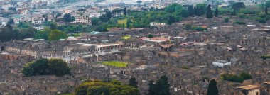 Pompei 'nin taştan binalar, sütunlar ve avlularla çevrili antik harabelerinin havadan görünüşü, yeşillik ve arka planda modern bir şehir manzarası..