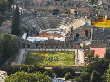 İtalya 'nın Pompei kentindeki Roma amfitiyatrosunun hava manzarası, ön planda harabeler, taş duvarlar, kemerler, yeşillikler ve modern sanat tesisleriyle çevrili..