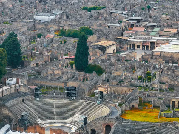 Pompei 'nin antik harabelerinin havadan görünüşü yarı dairesel taştan bir amfi tiyatro, çökmüş binalar, yollar ve selvi ağaçlı yeşillikler içeriyor..