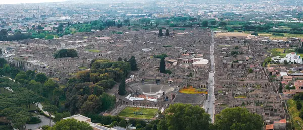 Pompei 'nin antik harabelerinin havadan görünüşü, yeşillik ve modern şehir manzarasıyla çerçevelenmiş bir Roma amfitiyatrosu, taş sokaklar ve binalar içeriyor..