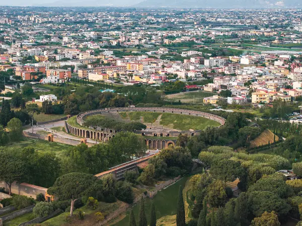 Yeşillik, modern binalar ve arka planda dağlık bir ufuk ile çevrili antik Pompei amfitiyatrosunun hava görüntüsü..