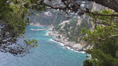 Capri 'nin turkuvaz suları, kayalık kayalıklar ve yeşil ağaç dalları tarafından çerçevelenmiş yamaç villalarıyla kıyı şeridi manzarası. Kamera daha fazla ayrıntı vermek için yansıtıyor.