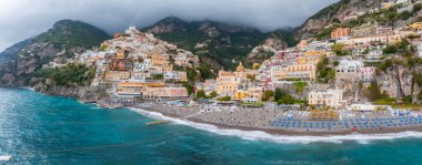 İtalya 'nın Amalfi Sahili' ndeki Positano manzarasında istiflenmiş binalar, kubbeli bir kilise, mavi şemsiyeli bir plaj ve turkuaz Akdeniz suları yer alıyor..