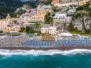 Positano 'nun tepelerdeki binaları, bir kilise kubbesi ve Amalfi kıyısındaki sahil boyunca mavi şemsiyeli bir plaj manzarası..
