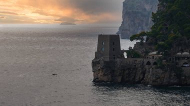 Positano, İtalya 'da bir sahil manzarası, kayalık bir uçurum üzerinde taş bir kule, çevresi yeşillik ve binalarla çevrili, gün batımında sakin suya bakan..