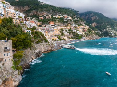 Sahil kenti Positano, İtalya, dik bir yamaçta istiflenmiş binalar, taş bir kule, turkuaz sular, botlar ve bulutlu bir gökyüzü..