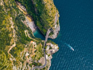 İtalya 'daki Amalfi Sahili' nin havadan görünüşü sarp kayalıkları, dolambaçlı bir yolu, turkuaz suları olan küçük bir sahili ve Tyrhenian Denizi 'nde bir tekneyi gösteriyor..