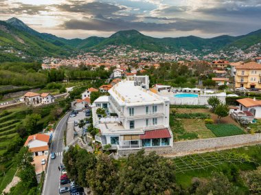 İtalya 'nın Amalfi kentindeki bir tepe manzarasında balkonları, havuzları, bahçeleri, terasotta çatıları ve yeşil tepeleri olan beyaz, lüks bir otel var..