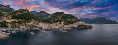 Amalfi, İtalya 'nın havadan görünüşü, kayalıklardaki renkli binalar, bir tepe oteli, tekneli bir liman, ve canlı pembe ve mor bir günbatımı gökyüzü..