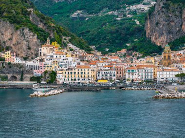 Amalfi, İtalya 'nın havadan görünüşü, renkli tepe binaları, önemli bir kilise çan kulesi, bir marina, kayalık dalgalar ve yemyeşil uçurumlar.