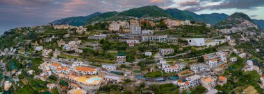 Teraslı manzaralar, turuncu çatılar, villalar, kiliseler, beyaz bir yapı ve dramatik bir gökyüzü altında yuvarlanan tepeler içeren Ravello, İtalya 'nın hava manzarası..